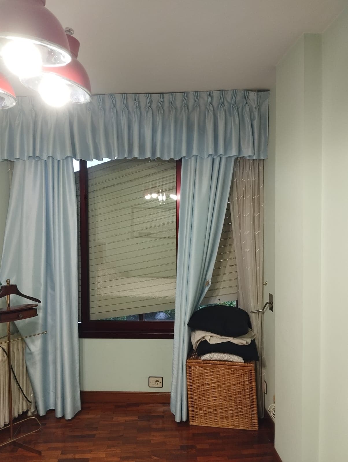 Dormitorio de una vivienda con cortinas azules y galería, y la persiana beige bajada a media altura tras la ventana de carpintería de madera oscura