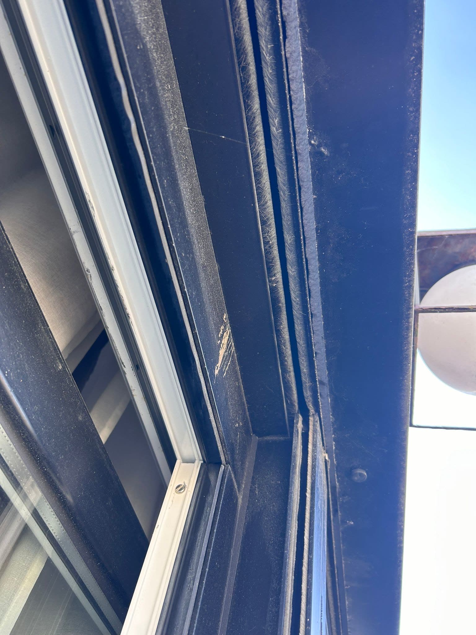Detalle del lateral de una ventana oscura visto desde fuera, con la guía de la persiana, el cepillo del burlete y la hoja entreabierta