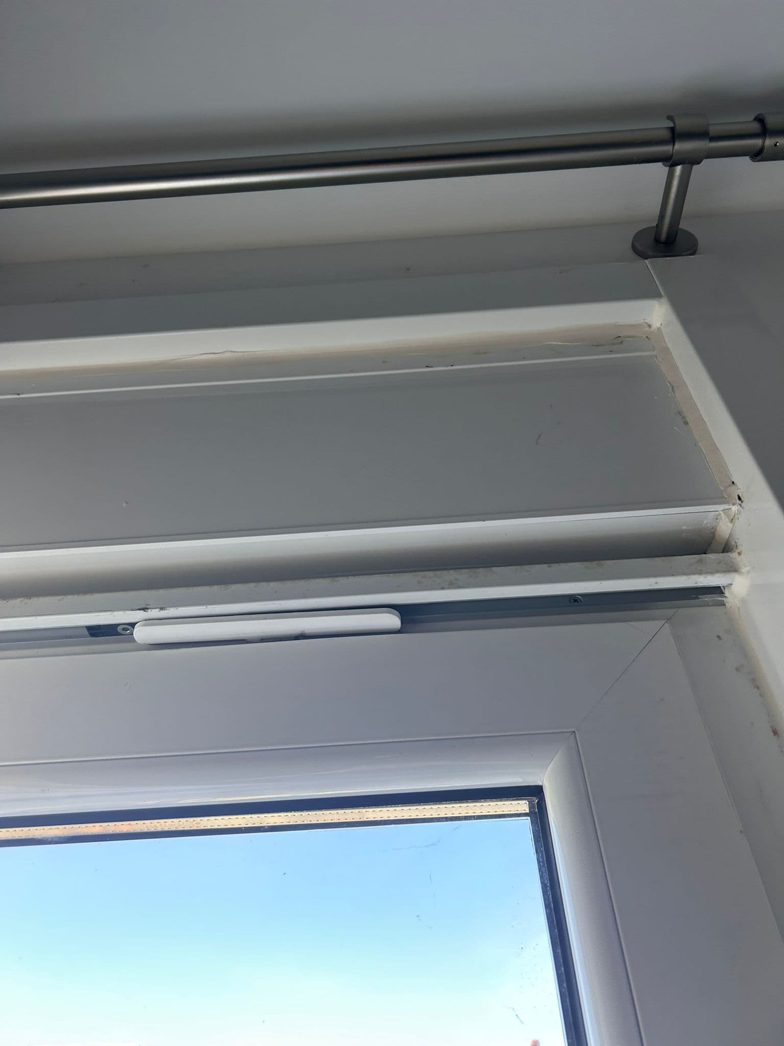 Esquina superior interior de una ventana blanca con el cajón de persiana y su tapa de registro, la barra de cortina justo encima y el carril con el soporte a la vista
