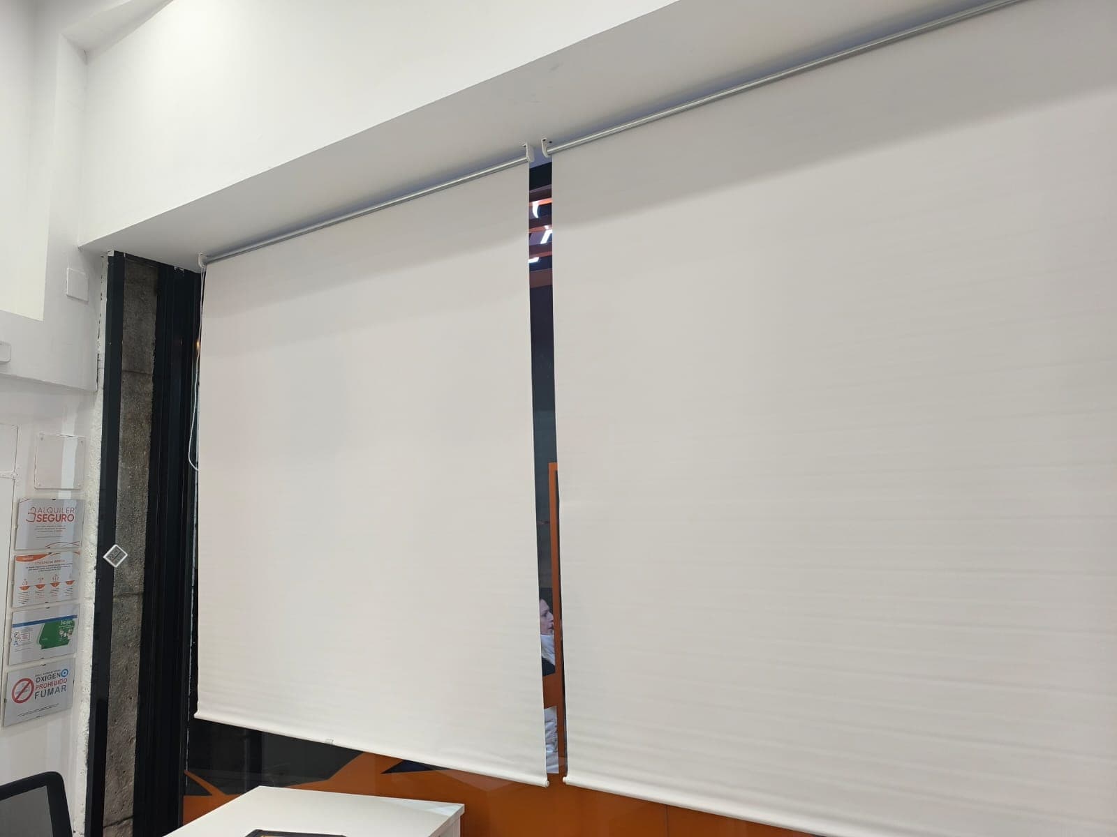 Dos estores enrollables blancos bajados sobre el escaparate de un bajo comercial, colgados de sus soportes a la pared junto a los carteles del local