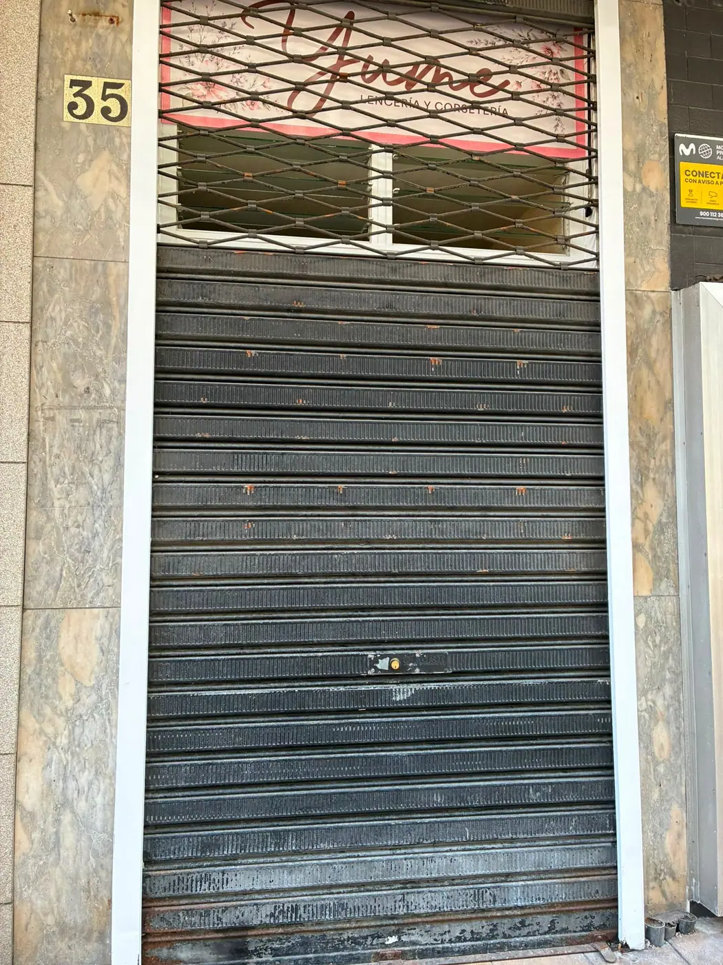 Cierre metálico enrollable bajado por completo en la entrada de un bajo comercial, con la cerradura centrada en la lama y una reja de ballesta sobre el escaparate